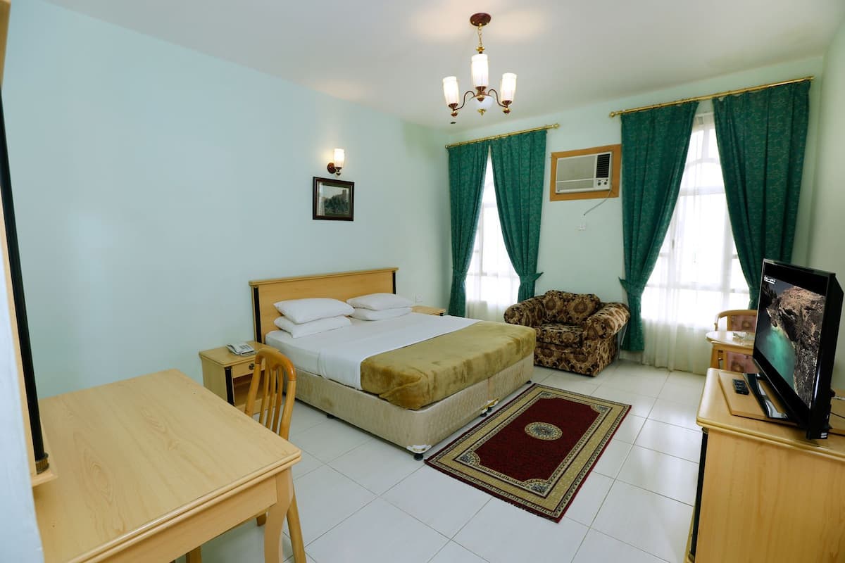 A bedroom with a bed, a desk, a chair, a TV, and a window with curtains.