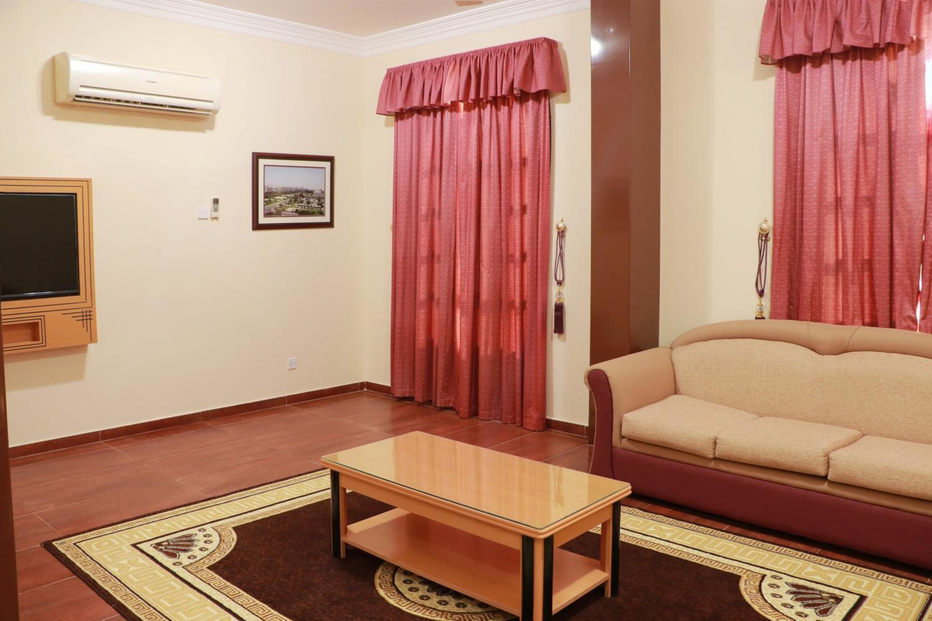 Una sala de estar con un sofá beige, una mesa de centro de madera, una cortina roja y un cuadro enmarcado en la pared.