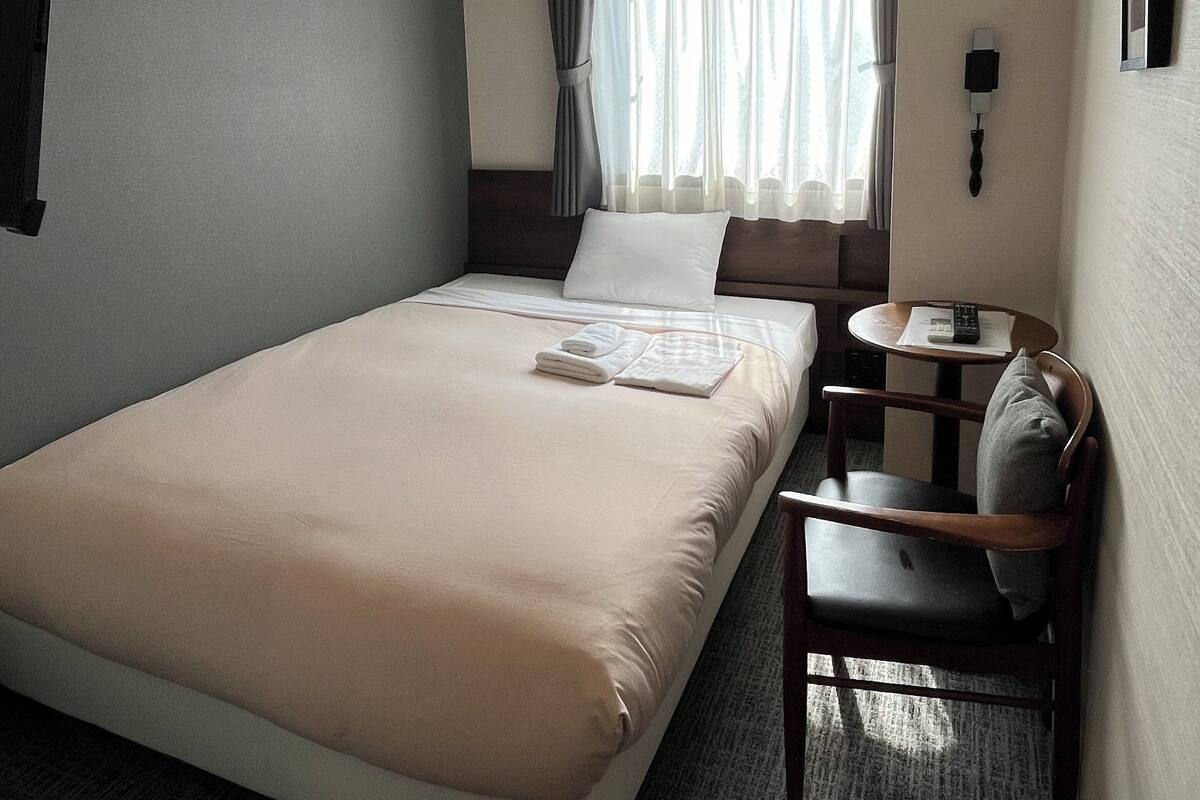 A hotel room with a single bed, a small table with a chair, a window with curtains, and a wall-mounted phone.
