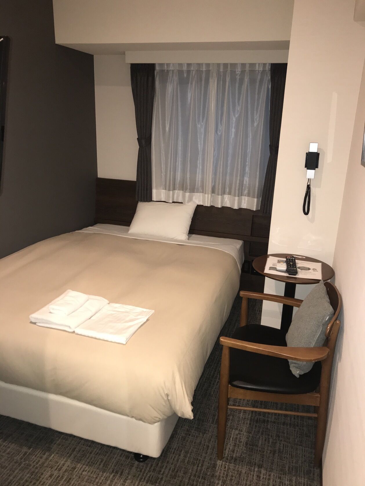 A hotel room with a bed, a chair, a small table with a remote control, and a phone on the wall.