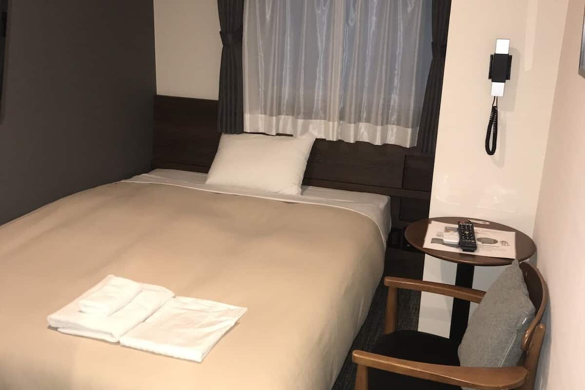 A hotel room with a bed, a chair, a small table with a remote control, and a phone on the wall.