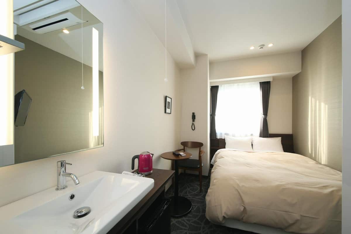 A hotel room with a large mirror, a white sink, a bed with beige bedding, a small round table with a chair, and a window with curtains.
