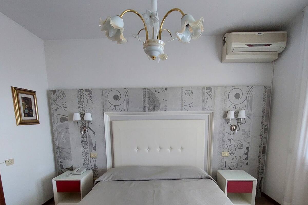 A bedroom with a bed, bedside tables, a chandelier, an air conditioner, and patterned wallpaper.