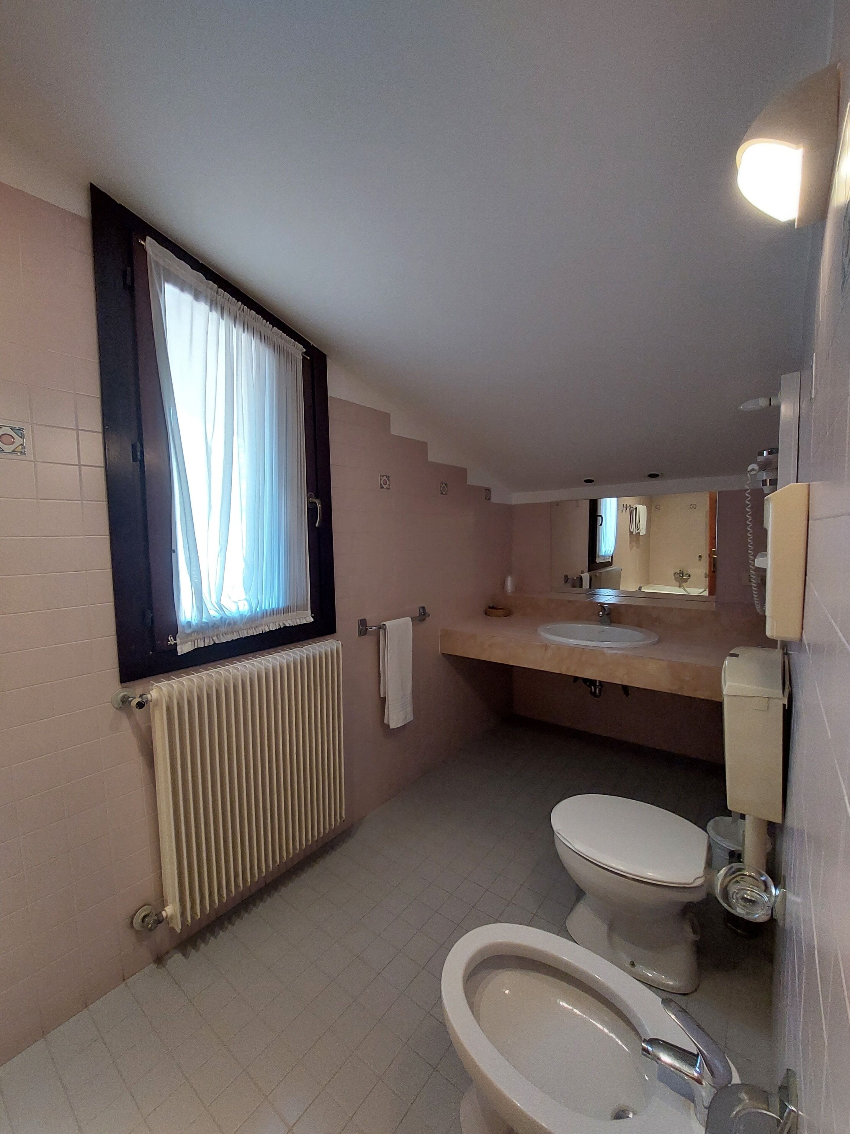 A small bathroom with a toilet, bidet, and sink. There is a window with curtains, a radiator, and a towel hanging on the wall.