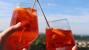 Dos vasos de bebida roja con pajita, sostenidos por manos, con un cielo despejado y un paisaje lejano al fondo.