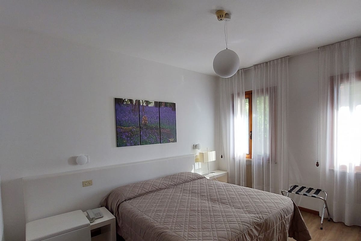 A bedroom with a large bed, a bedside table with a lamp, a small bedside table with a phone, a radiator, and a window with curtains.