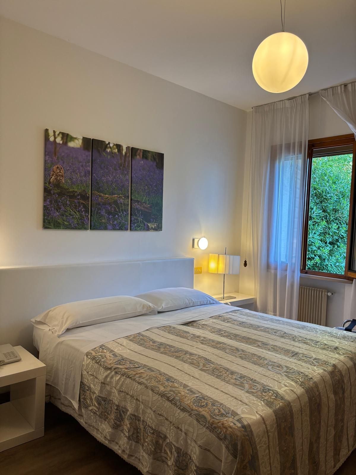 Een slaapkamer met een bed, een nachtkastje, een lamp, een raam met gordijnen en een kunstwerk aan de muur.
