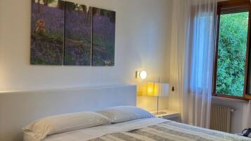 Een slaapkamer met een bed, een nachtkastje, een lamp, een raam met gordijnen en een kunstwerk aan de muur.