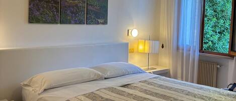 A bedroom with a bed, a bedside table, a lamp, a window with curtains, and a wall-mounted artwork.