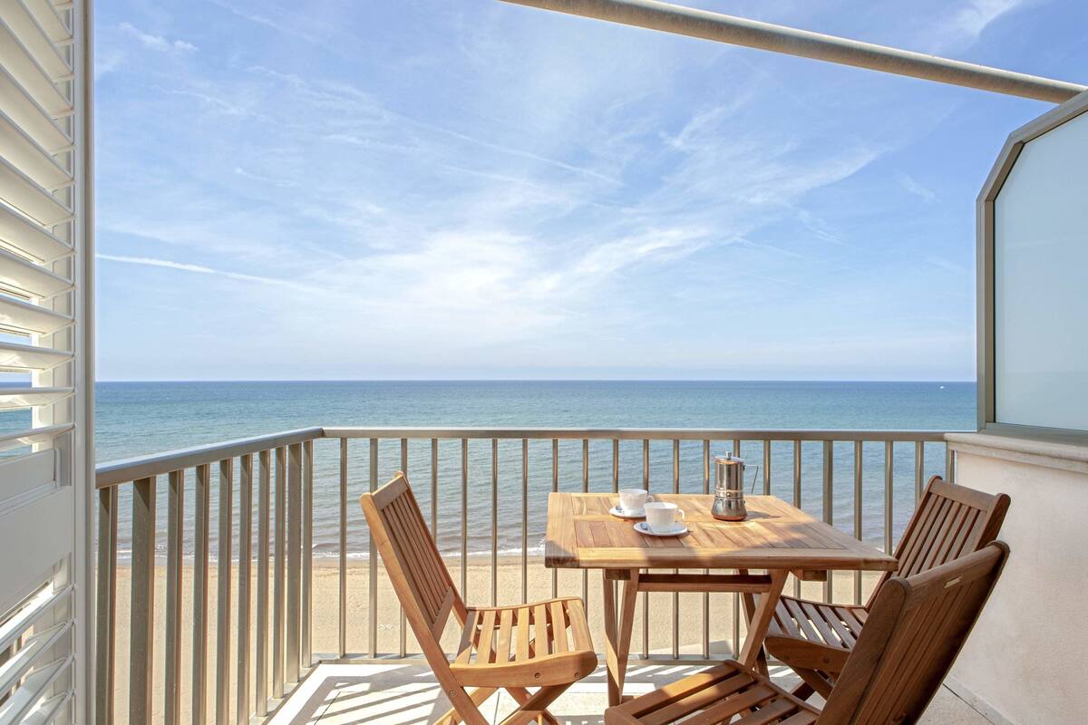 Un balcón con mesa y sillas de madera, con vista a una playa y al mar.