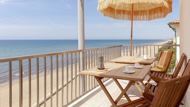 Un balcon avec vue sur la plage et l’océan, des meubles en bois et un parasol en paille.