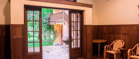 Un interior de madera con un amplio ventanal abierto a un patio, una mesa redonda pequeña y dos sillas de mimbre.