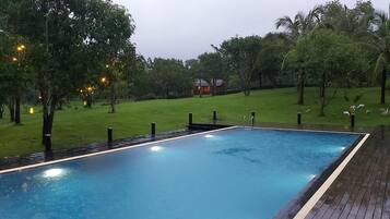 Una piscina rectangular con un borde de madera, con árboles y una colina cubierta de hierba al fondo.