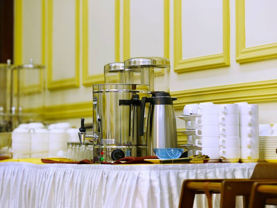 Una mesa de buffet con una cafetera, dispensadores de condimentos y vasos blancos apilados.