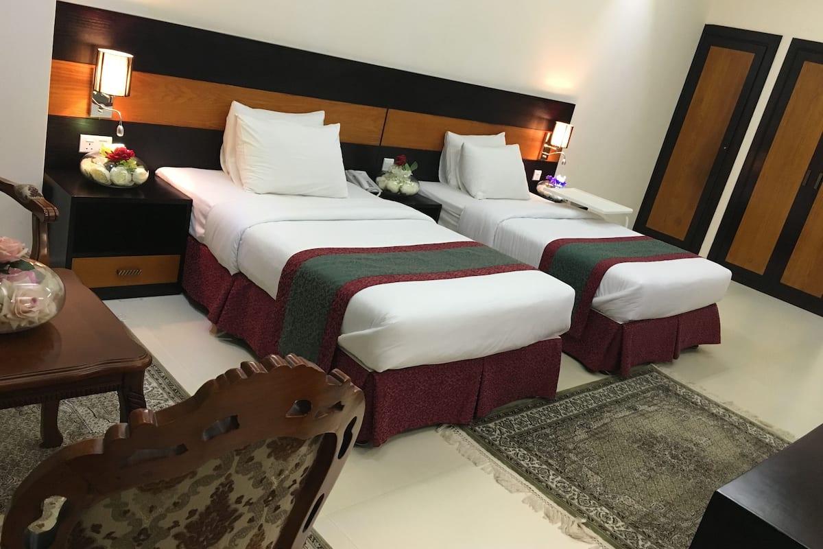 A hotel room with two single beds, a nightstand with a lamp, a small table with a vase of flowers, a chair, and a rug.