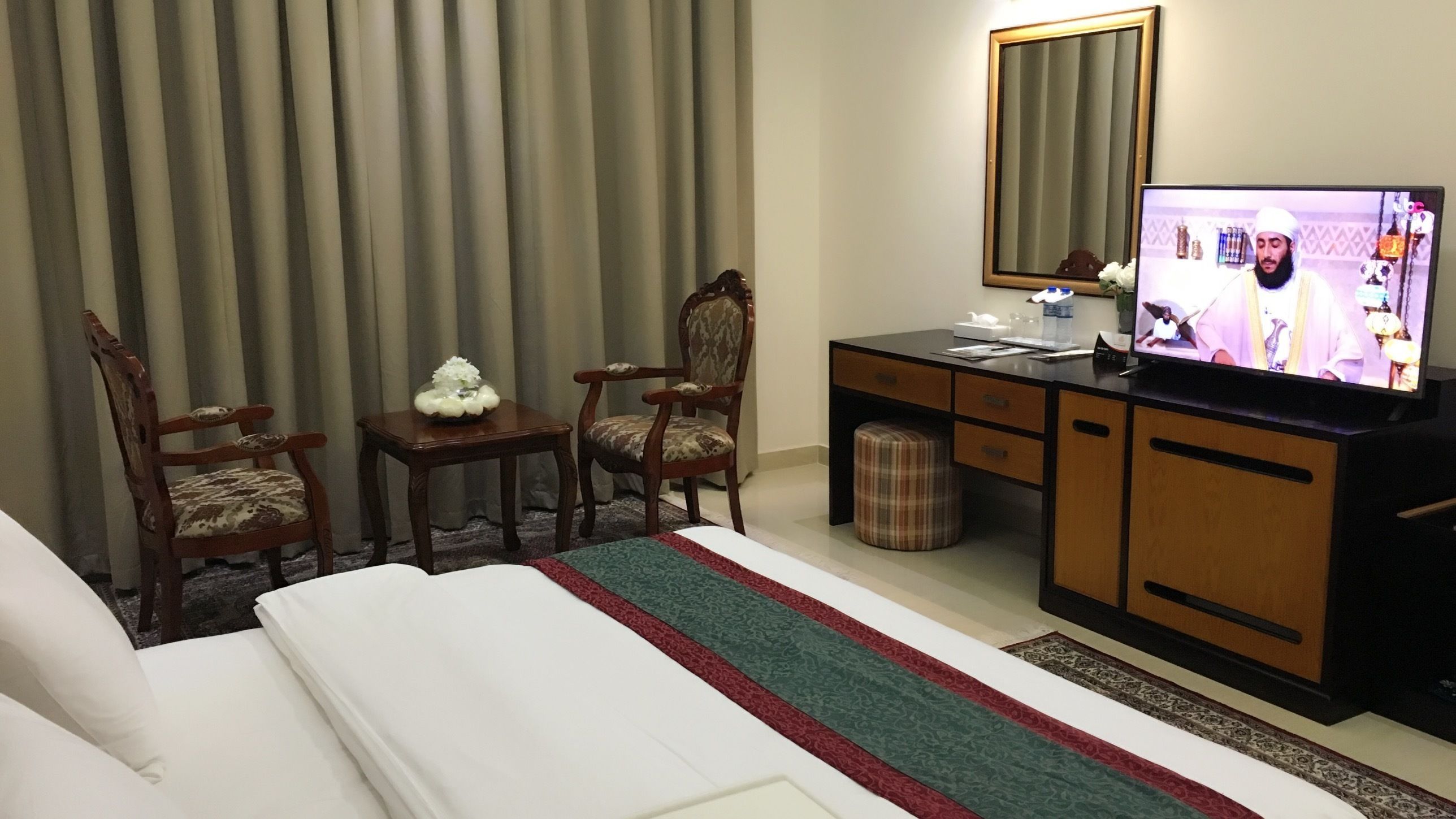A hotel room with a bed, a desk with a computer, two chairs, a small table with a flower arrangement, a mirror, and a television.