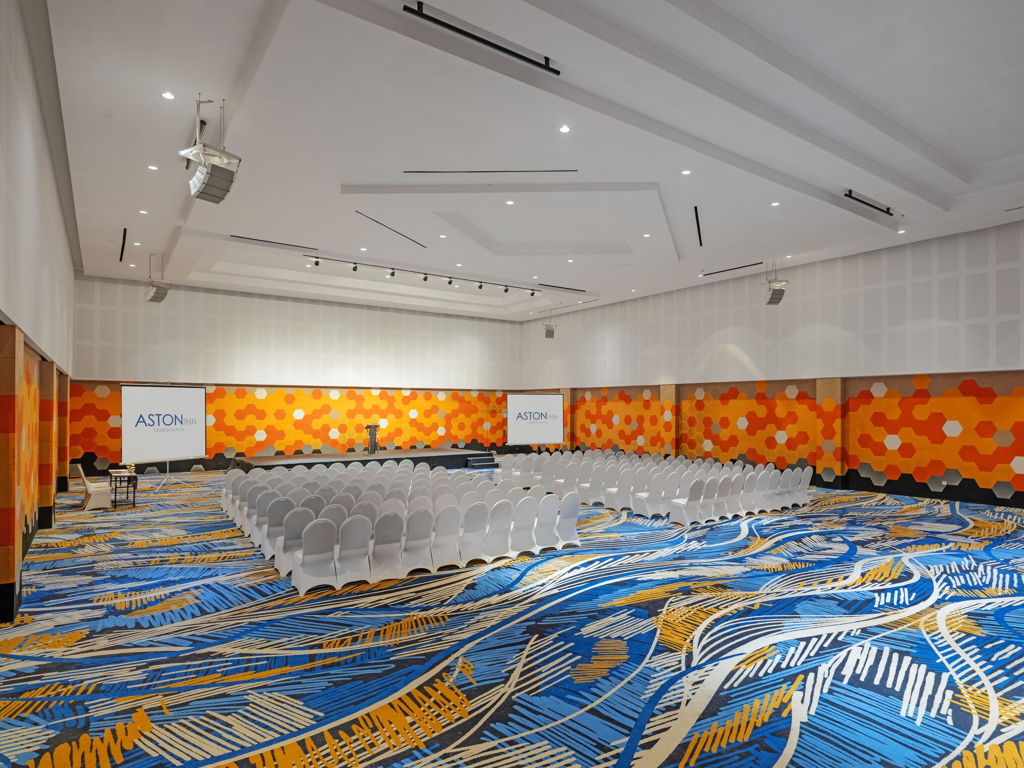 A large conference room with rows of white chairs, a stage with a screen, and a colorful carpet.