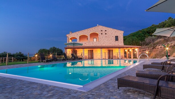 A swimming pool with a stone deck, a building with arched windows, and outdoor furniture. - CasalFarano (Cupra Marittima)