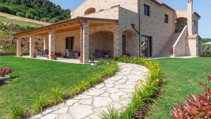 A two-story house with a covered patio, stone pathway, and a garden with flowers and plants. - CasalFarano (Cupra Marittima)