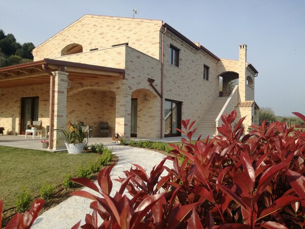A two-story house with a brick exterior, a covered patio, and a well-maintained garden with red-leafed plants. - CasalFarano (Cupra Marittima)
