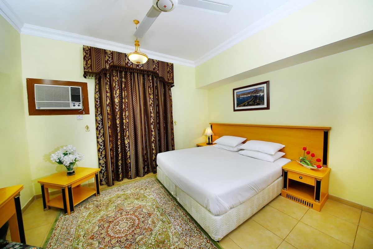 A hotel room with a bed, a nightstand, a desk, a chair, a window with curtains, a ceiling fan, an air conditioning unit, and a framed picture on the wall.