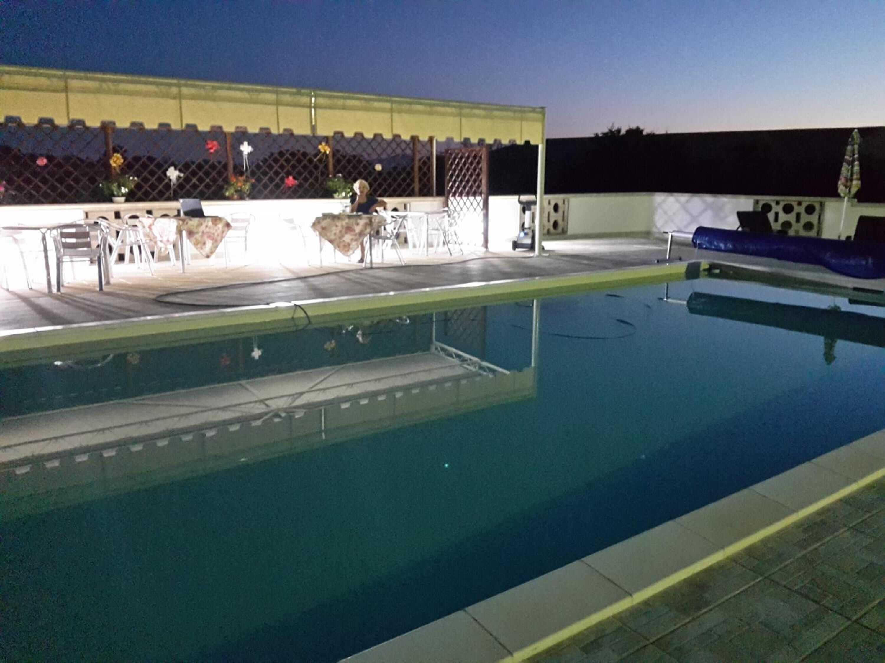 A swimming pool with a sunlit deck area, a covered seating area, and a fenced enclosure with plants.