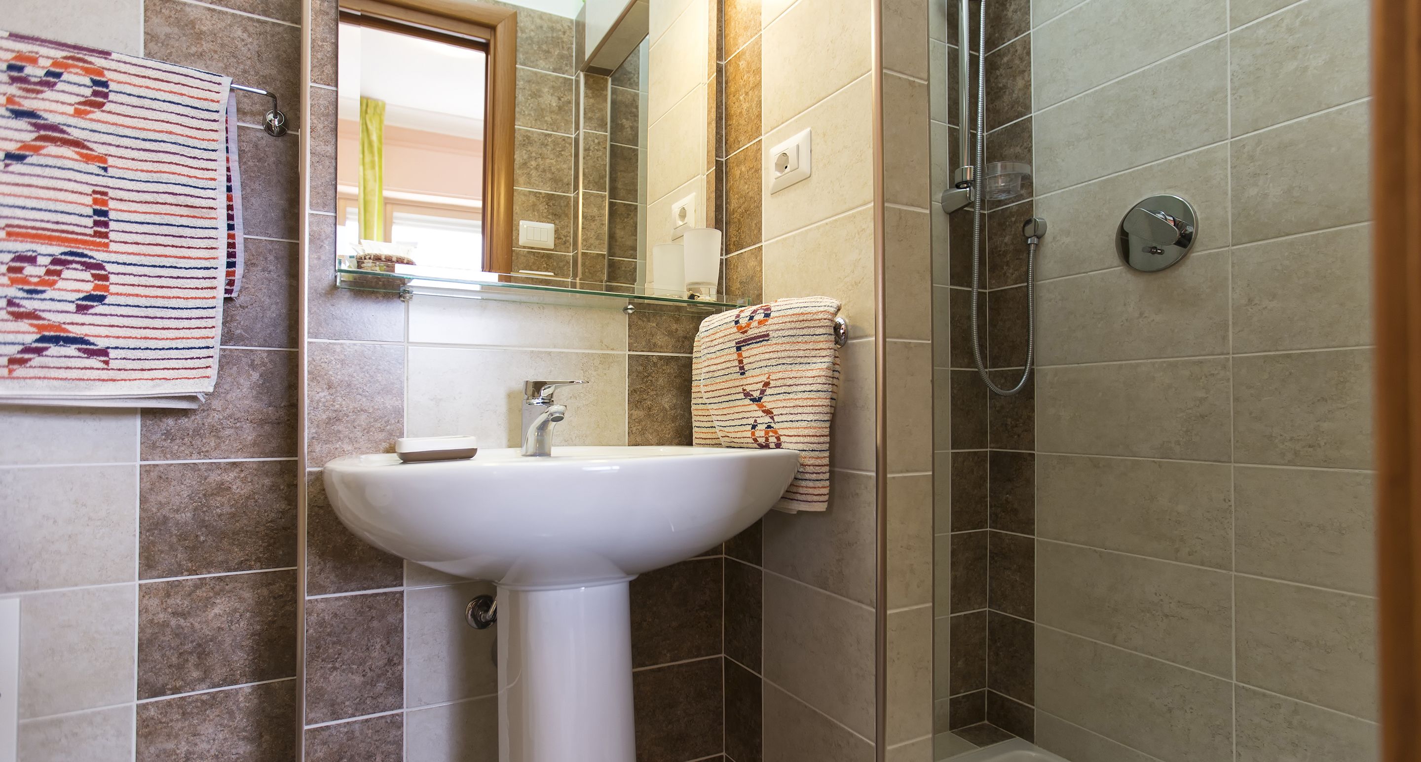 A bathroom with a white sink, a glass shelf above it, a towel rack with a striped towel, a mirror, and a shower area.