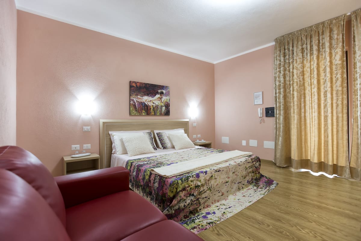 A hotel room with a bed, a nightstand, a red chair, a painting on the wall, and a window with curtains.