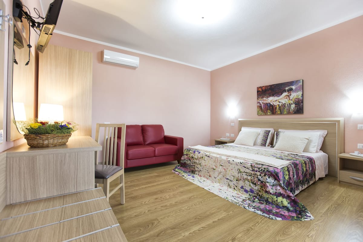 A bedroom with a bed, a red sofa, a desk with a lamp, and a painting on the wall.