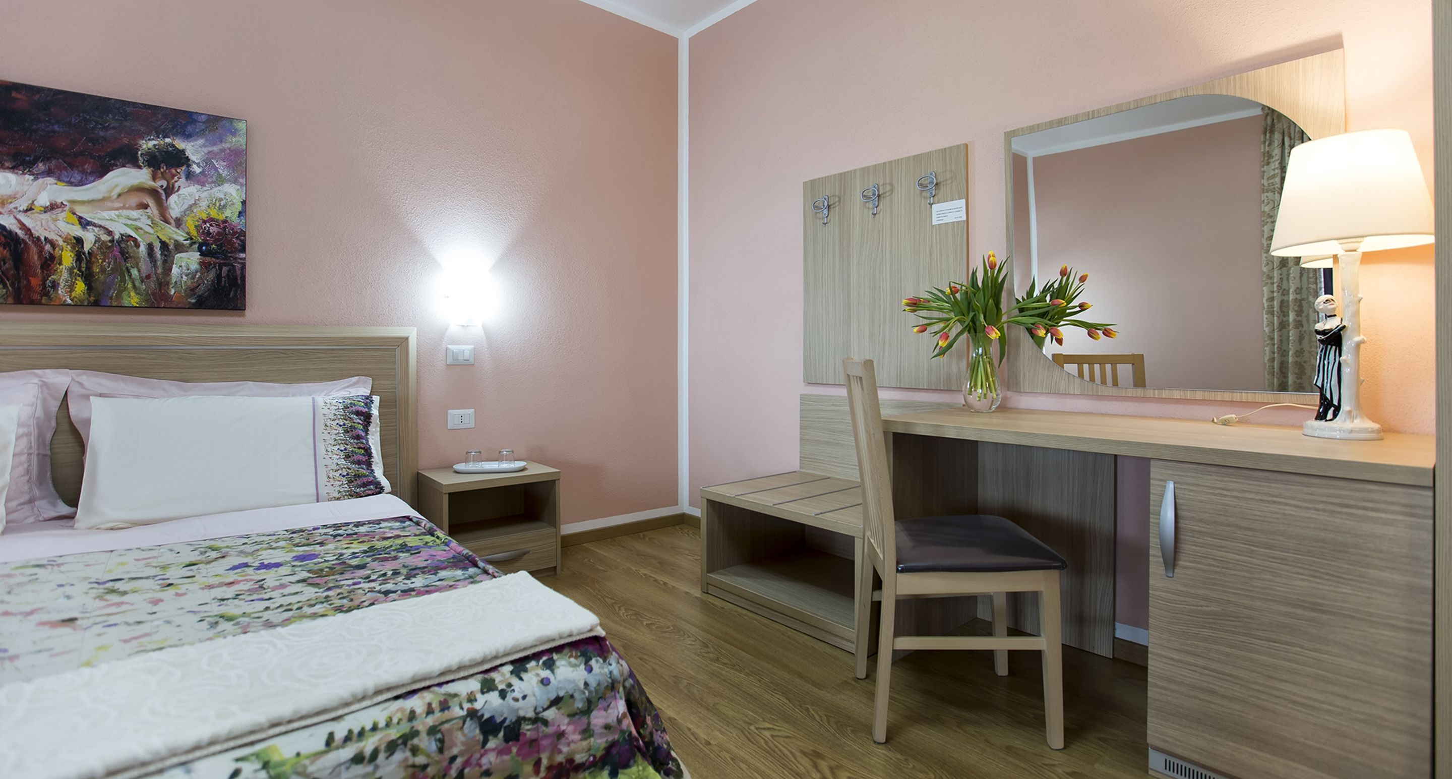 A hotel room with a bed, a desk with a chair, a mirror, a vase with flowers, and a painting on the wall.