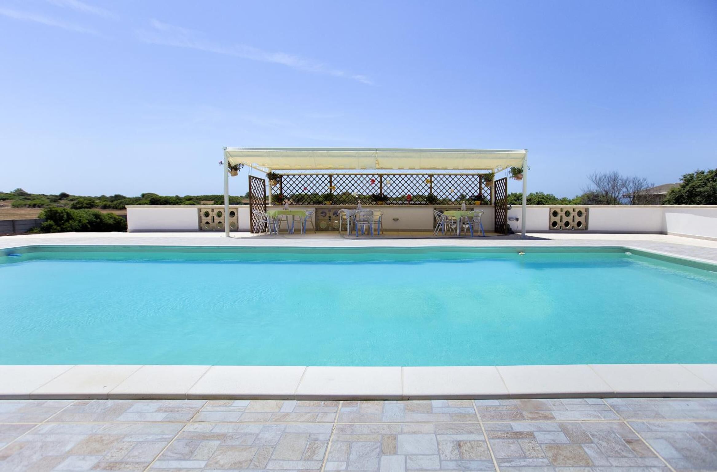 Una piscina con una zona de estar cubierta y sillas, rodeada por un muro bajo y un cielo despejado.
