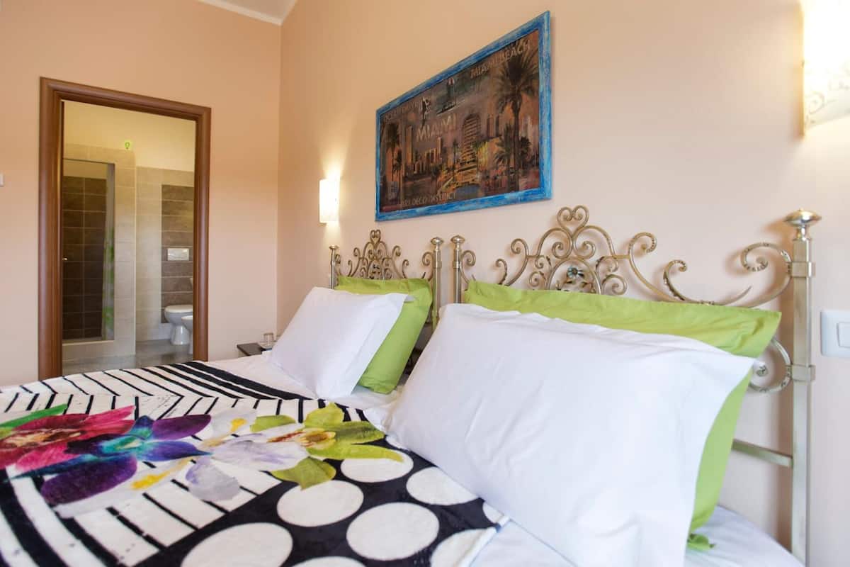 A double bed with green and white pillows, a black and white polka dot bedspread, and a framed picture of Miami on the wall.