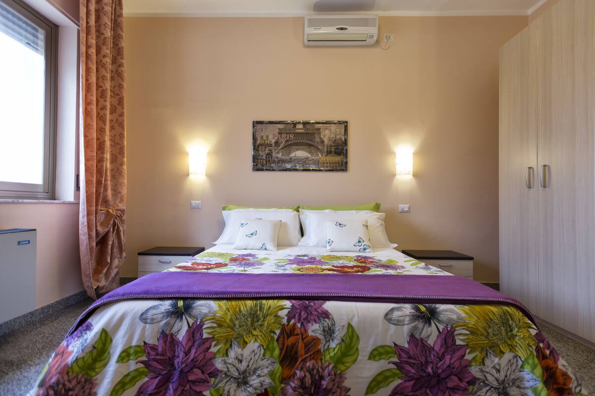A bedroom with a bed, a nightstand, a wardrobe, a window with curtains, and a wall-mounted air conditioner.
