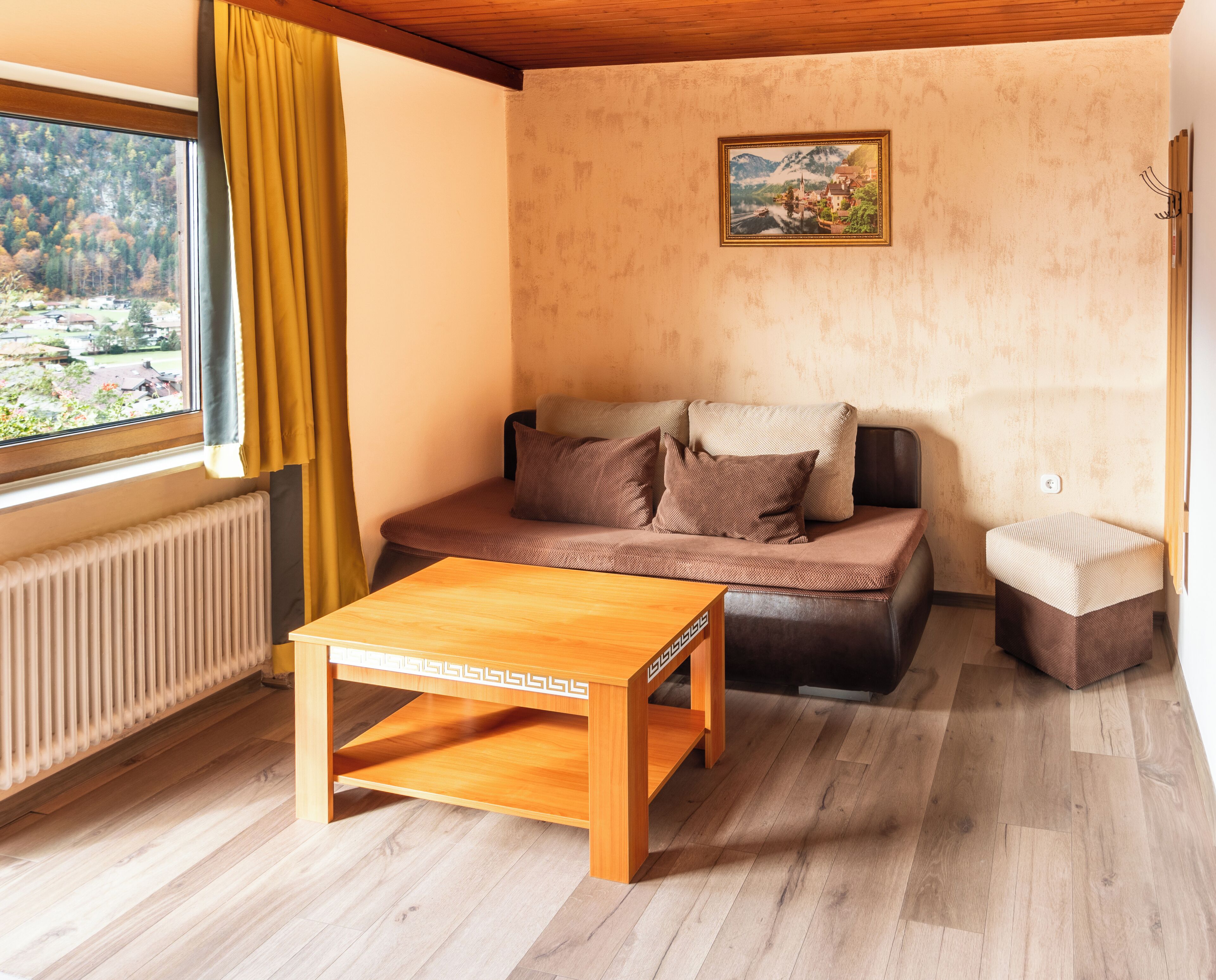 A cozy living room with a wooden table, a sofa with cushions, a radiator, and a window with a view of trees and mountains.