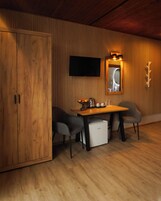 A wooden-paneled room with a wooden table, two chairs, a small white refrigerator, a television mounted on the wall, and a mirror with a light above it.