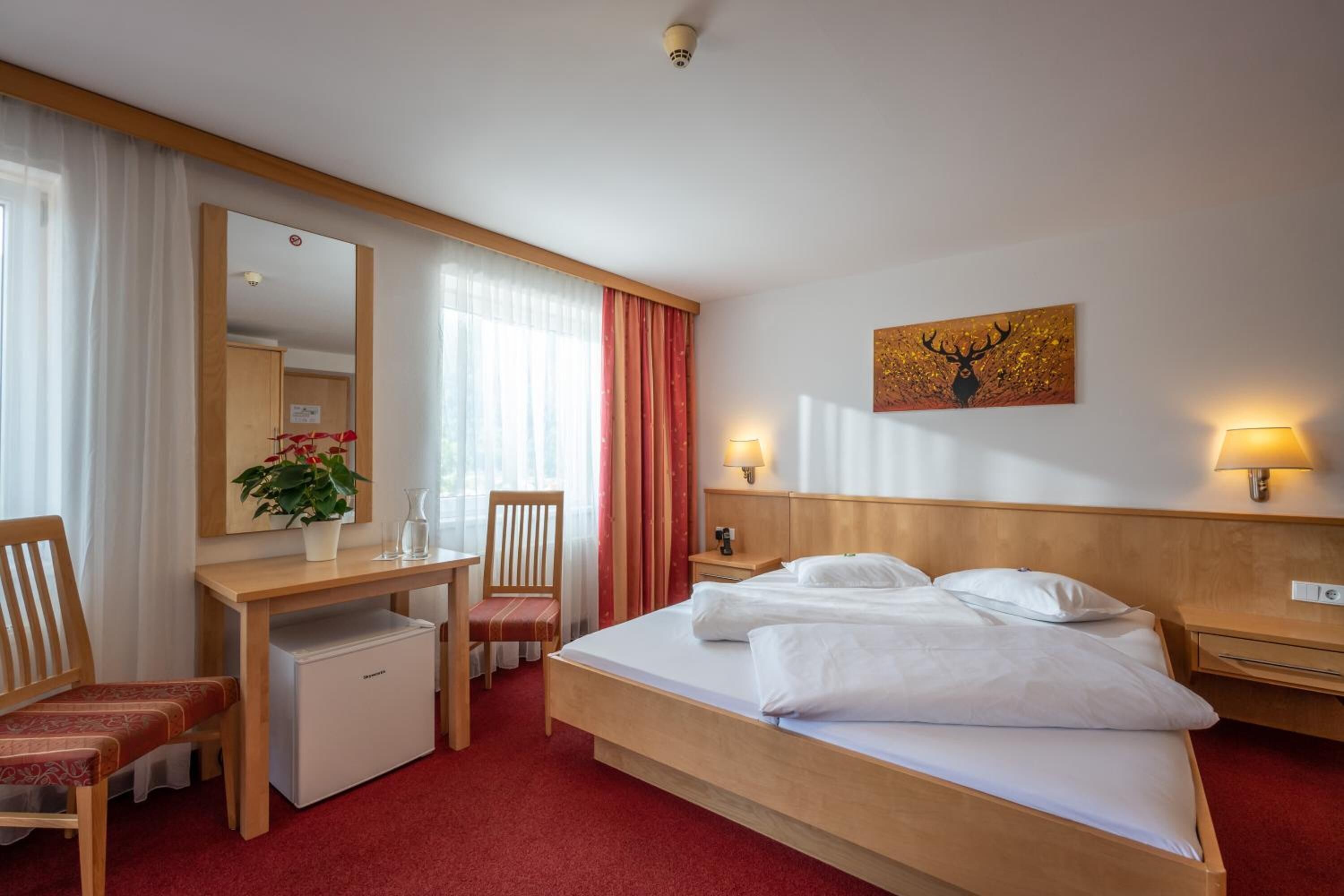 Quarto de hotel com cama, escrivaninha, cadeira, pequena geladeira, espelho, luminária e um quadro na parede.
