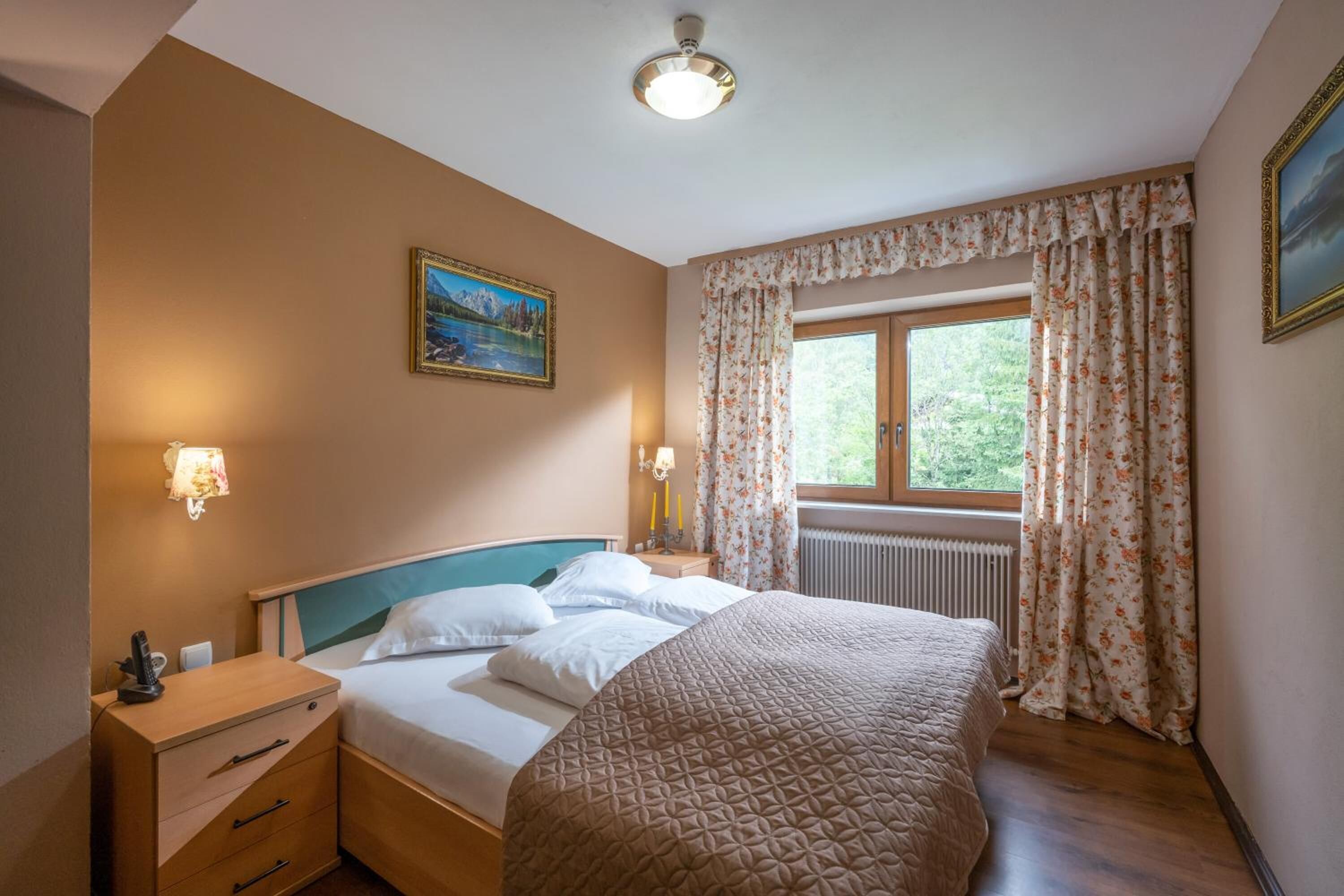 A bedroom with a bed, bedside lamps, a nightstand, a dresser, a window with curtains, and a painting on the wall.