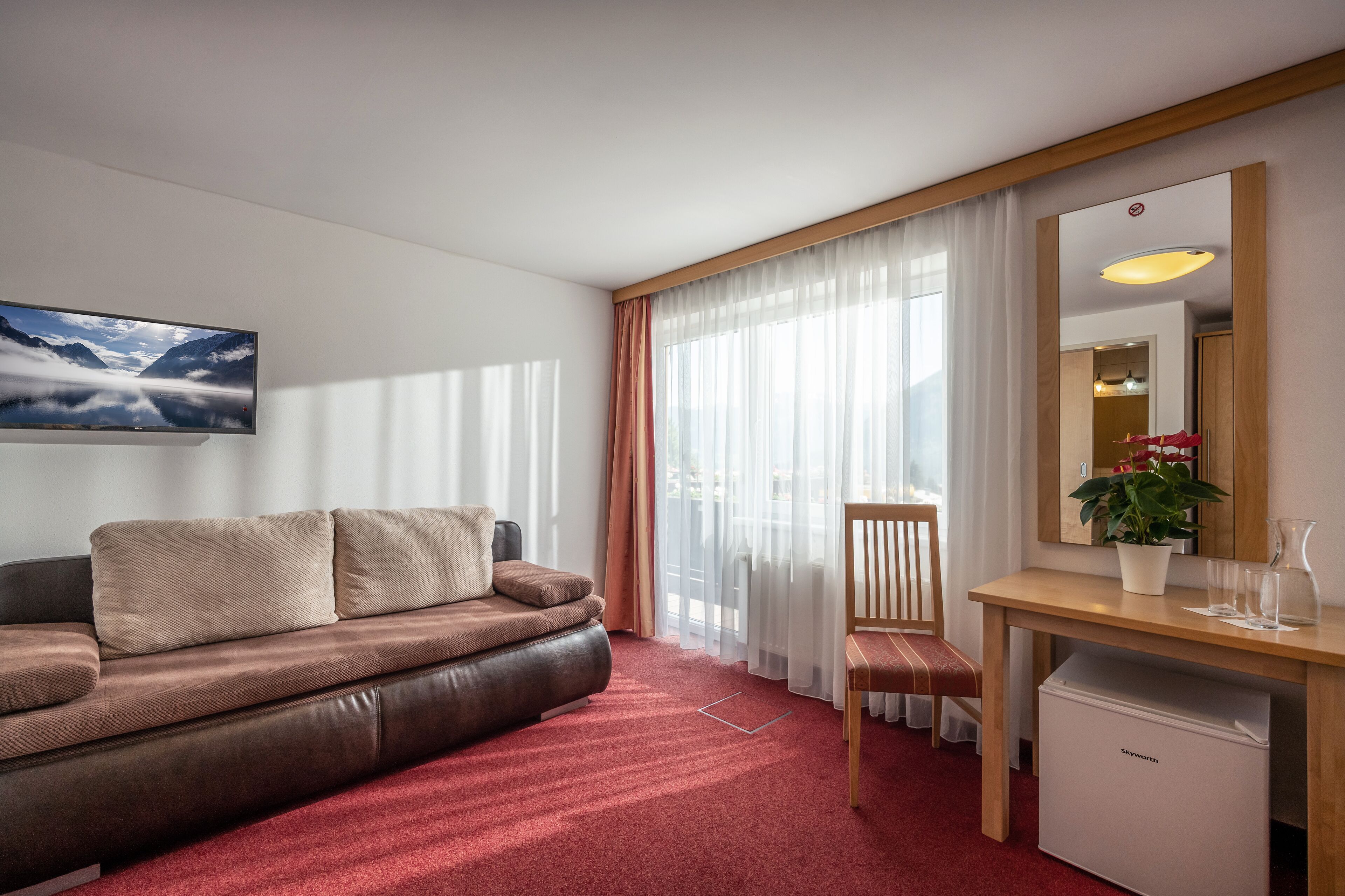 A modern hotel room with a leather sofa, a wooden desk, a small refrigerator, and a large window with sheer curtains.