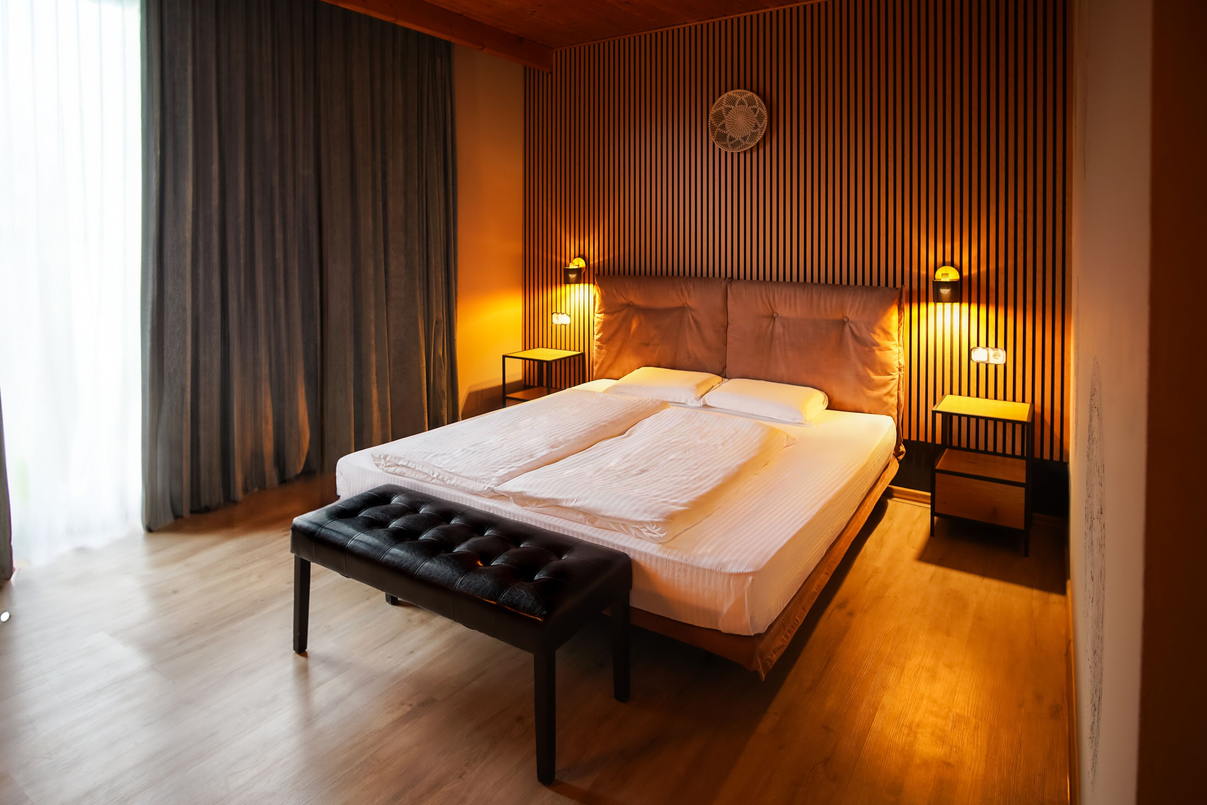 Un dormitorio moderno con una cama grande, dos mesitas de noche con lámparas, un banco y un diseño de pared con listones de madera.