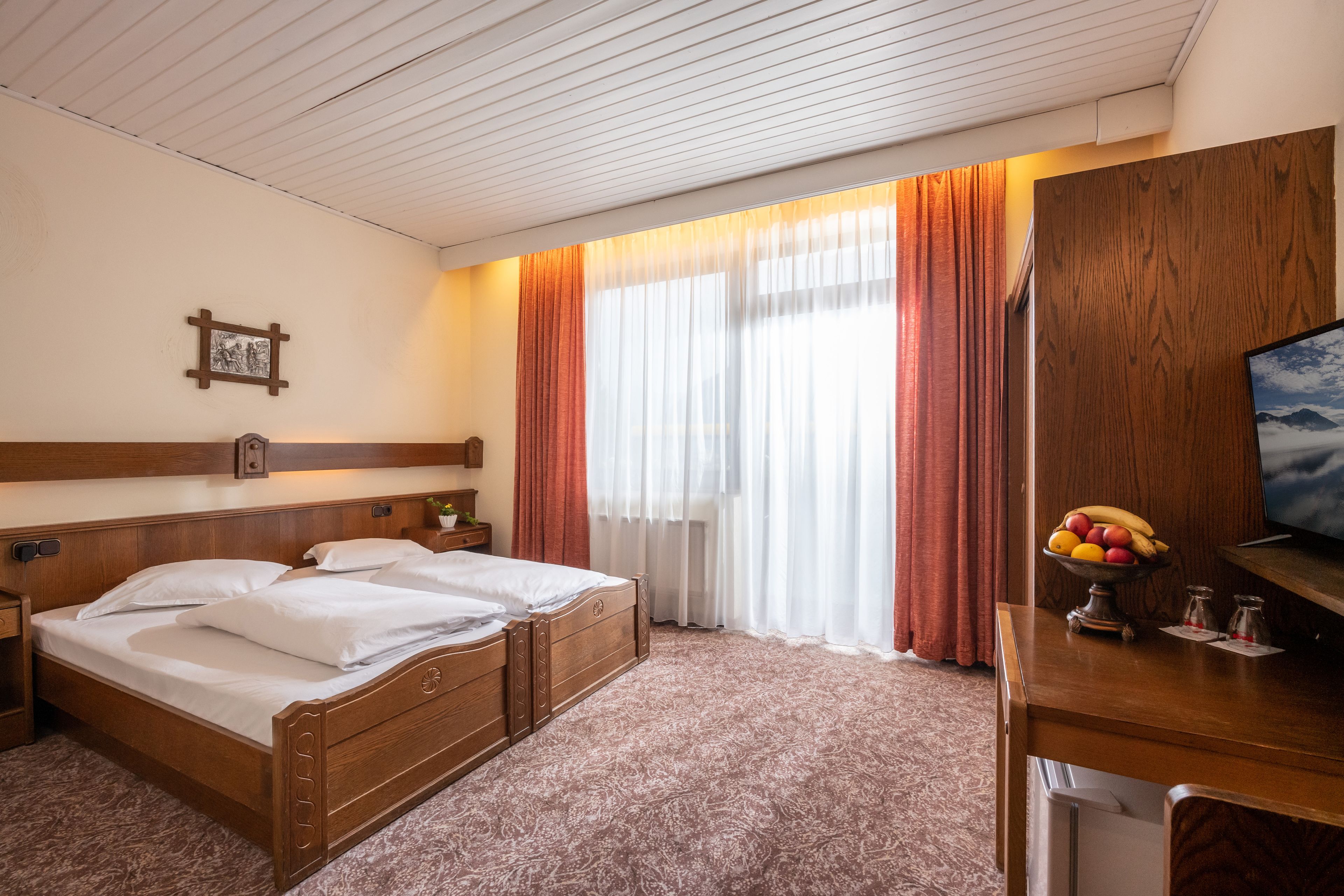 Une chambre d’hôtel avec un lit double, une tête de lit en bois, un bureau avec un panier de fruits et un téléviseur.