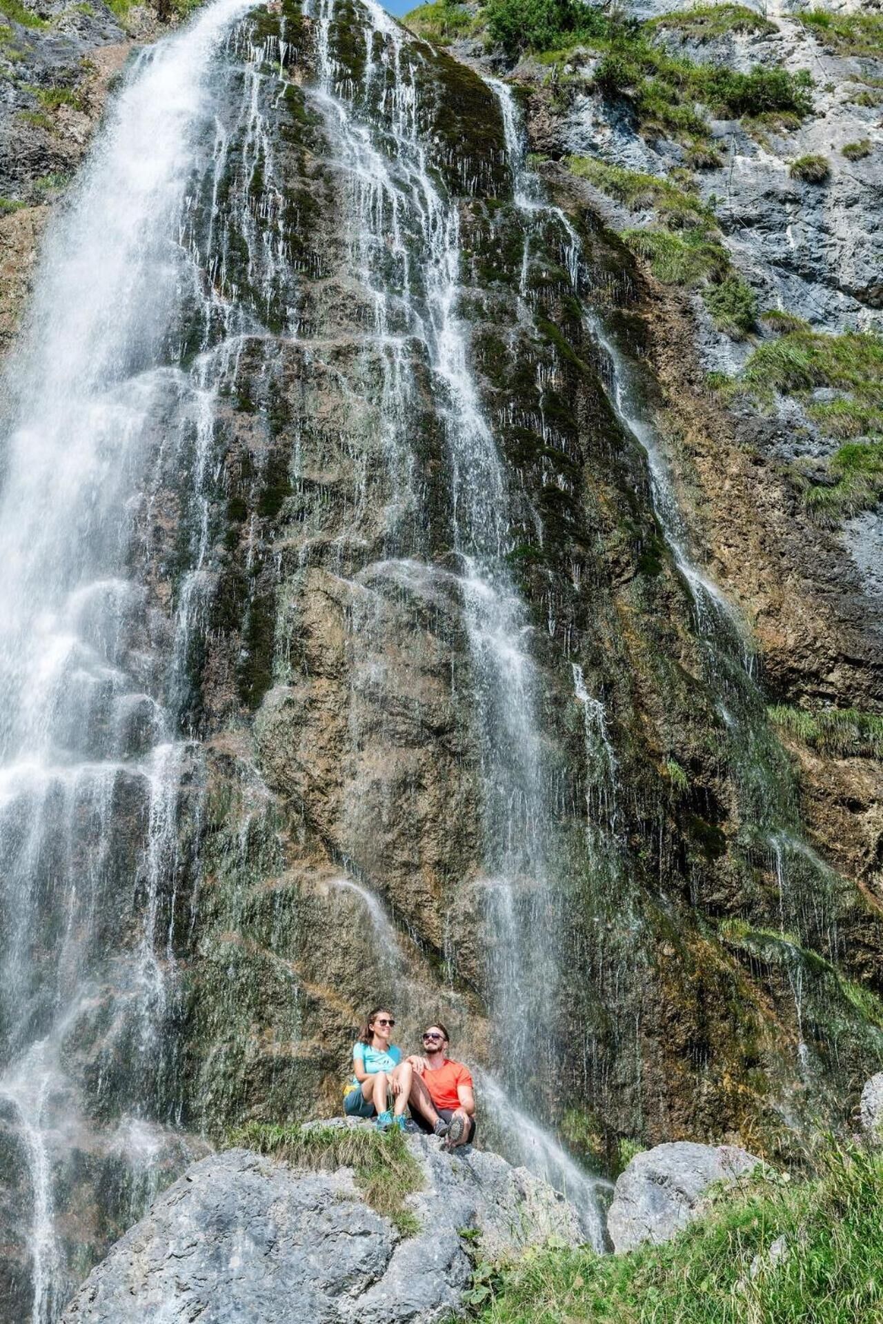 Ein Wasserfall stürzt über eine felsige Klippe, darunter sitzen ein Paar auf einer Felsstufe.