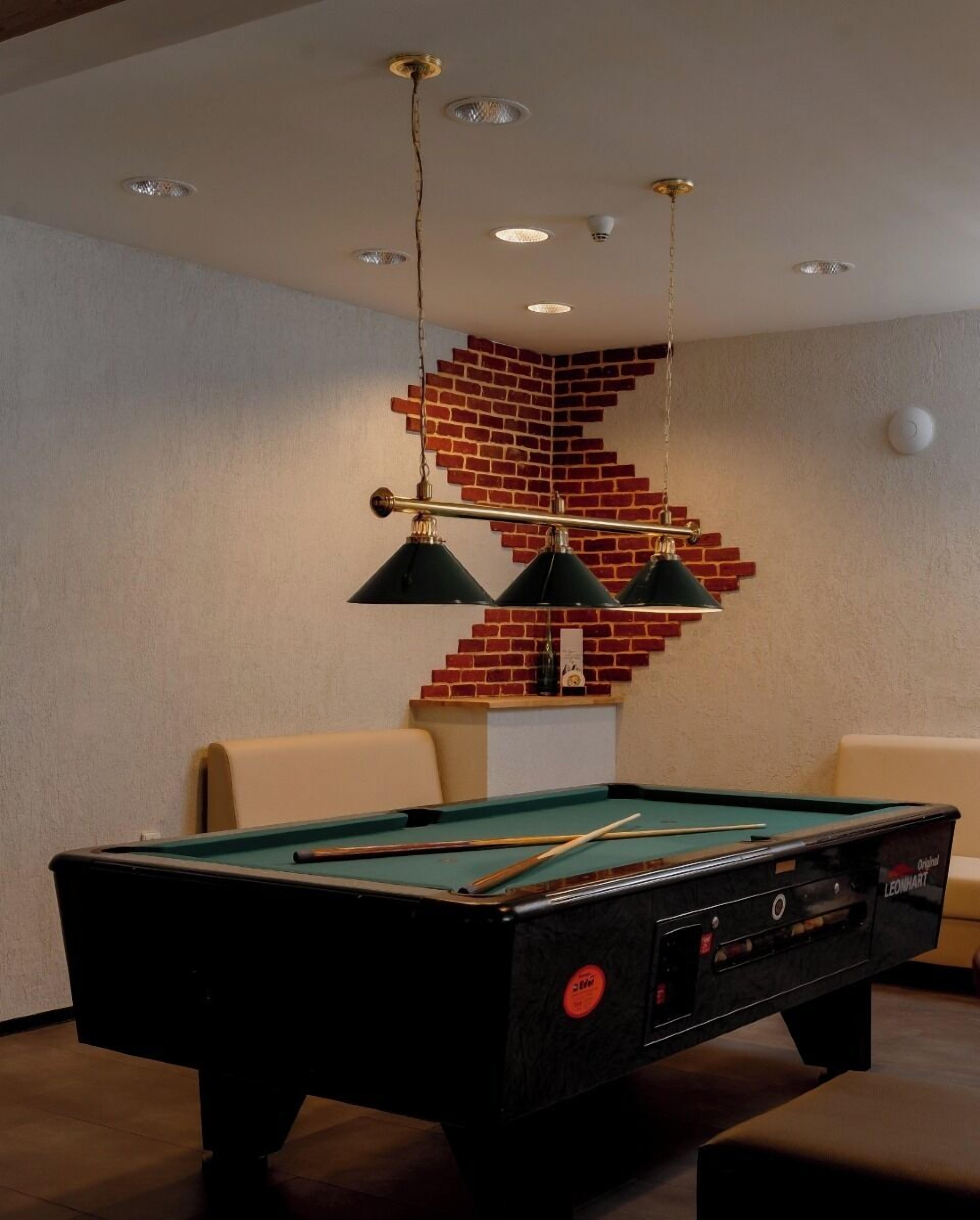 Een pooltafel met een groen vilt oppervlak, omringd door beiges zitzakken. Drie zwarte hanglampen zijn aan het plafond bevestigd, en er is een muur met een bakstenen patroon achter de zitzakken.