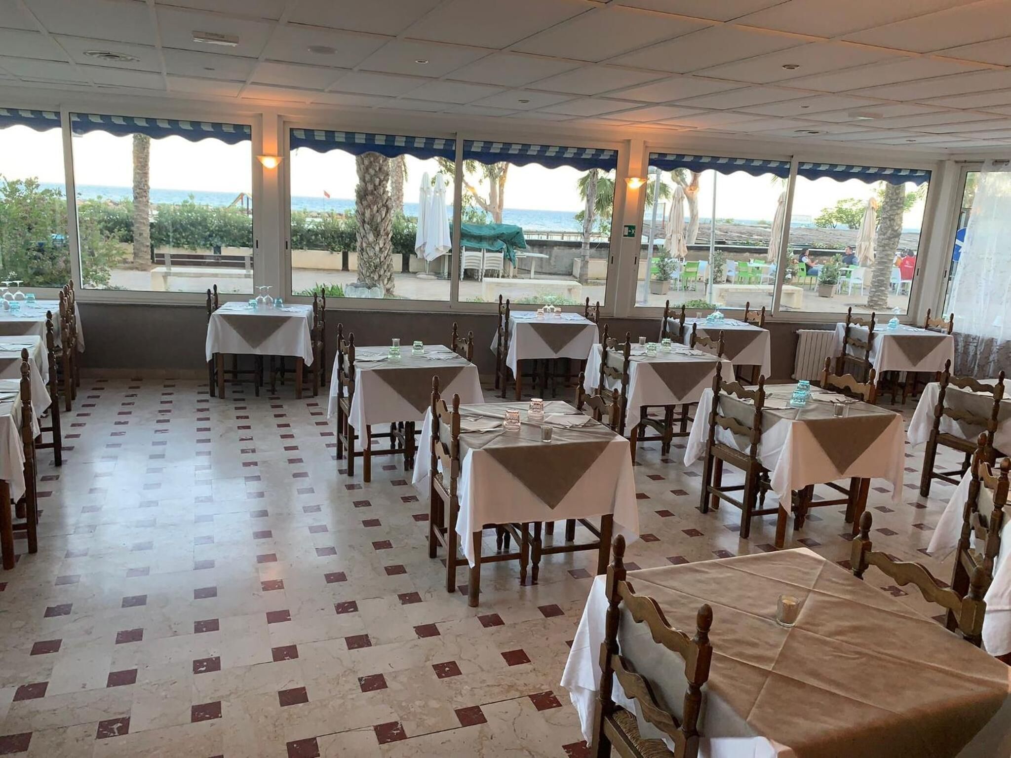 Ein Restaurant mit Tischen und Stühlen, großen Fenstern mit Blick ins Freie und einem gefliesten Boden.