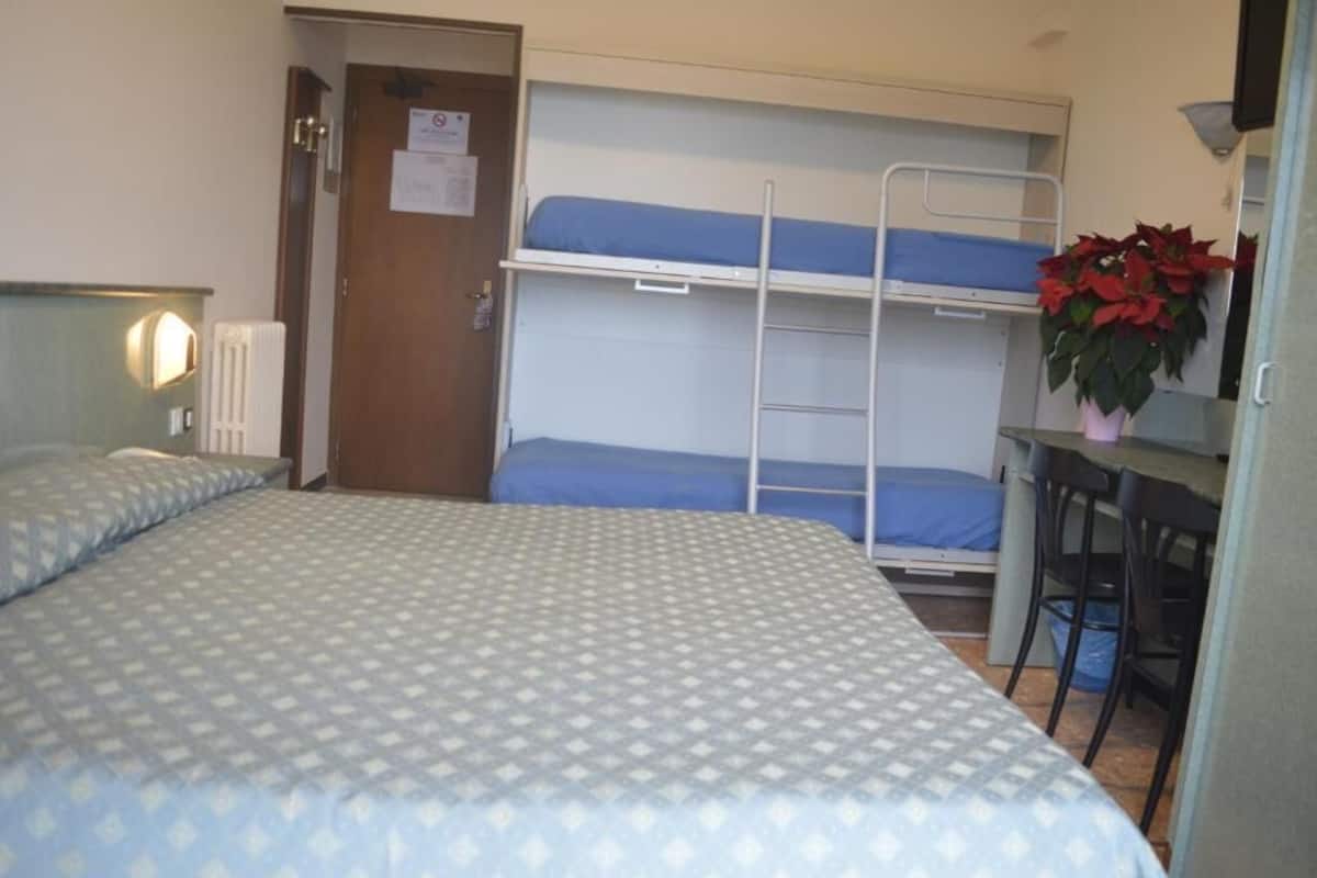 Une petite pièce comprenant un lit superposé, un bureau avec deux chaises, un radiateur et une porte sur laquelle est affiché un avis.