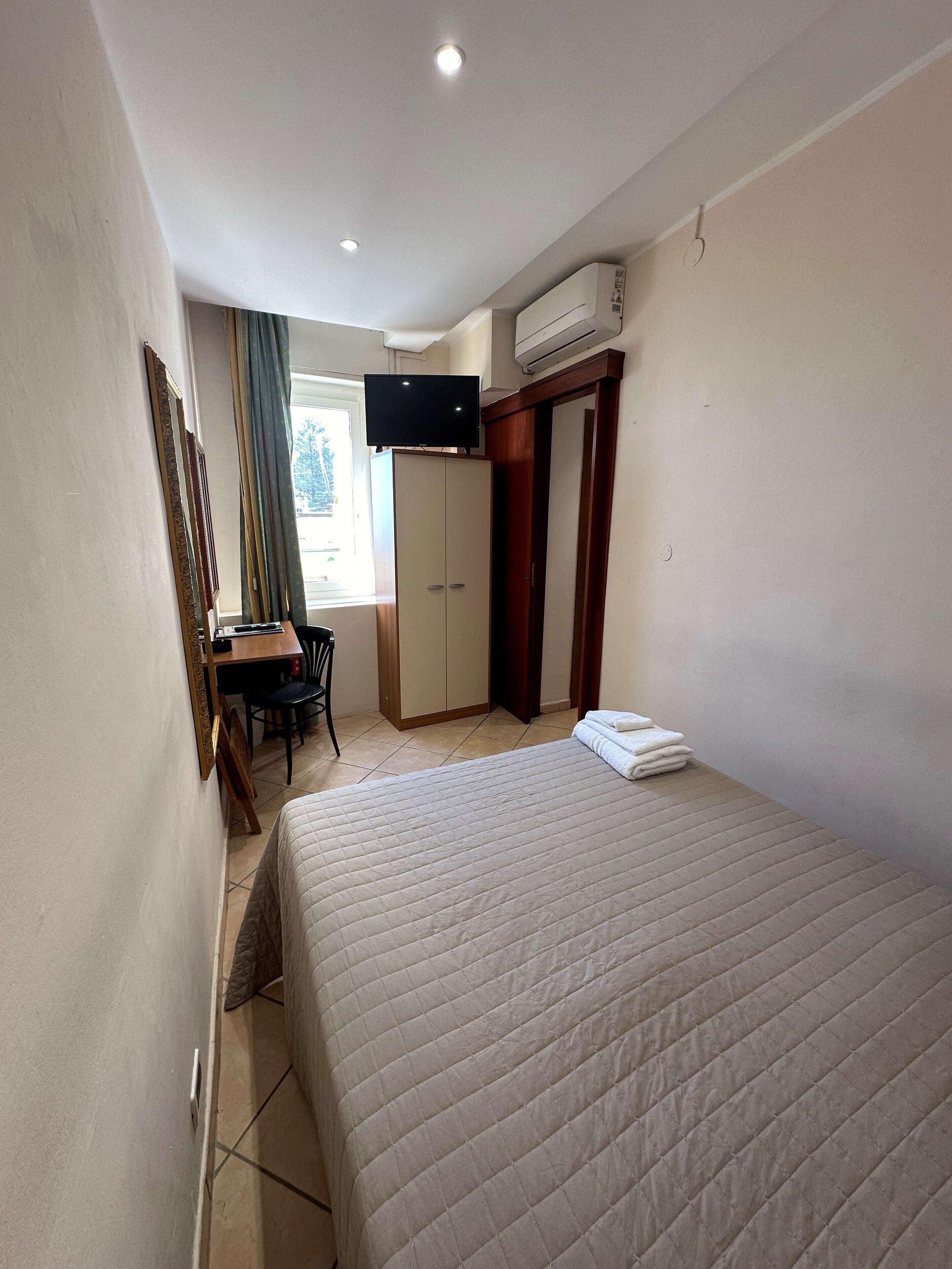A hotel room with a bed, a desk with a chair, a television, and an air conditioning unit.
