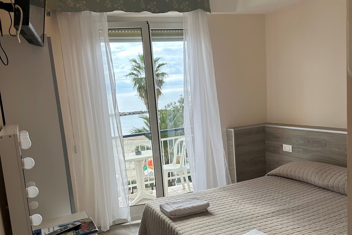 Une chambre d’hôtel comprenant un lit, une coiffeuse avec éclairage, une chaise, un bureau avec un téléphone et une vue sur la mer par la fenêtre.