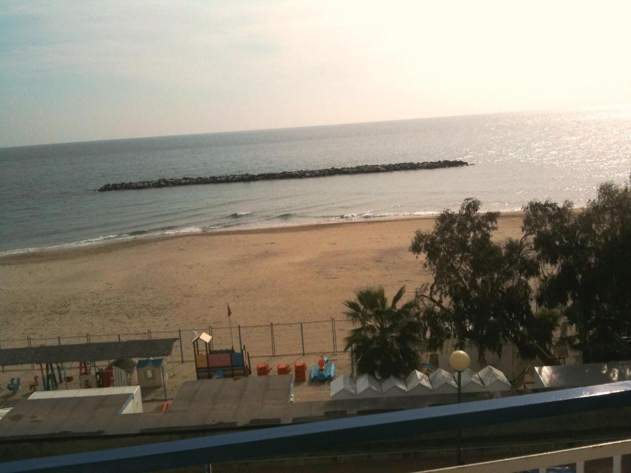 A beach with a long jetty extending into the water, a sandy shore, and some beach facilities including a small shelter and a fence.