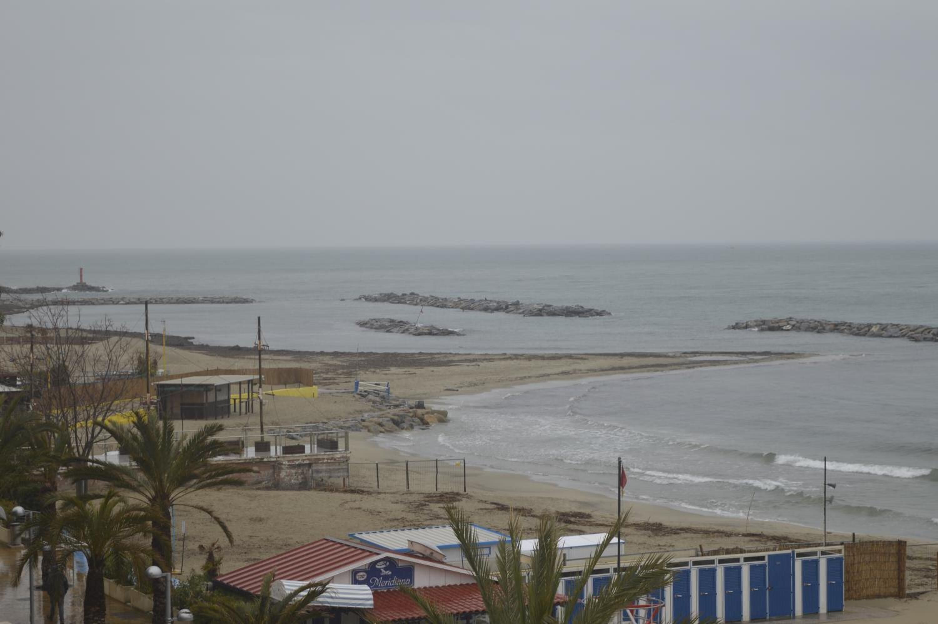 A beach view with a rocky breakwater, a lifeguard tower, and a row of blue beach huts.