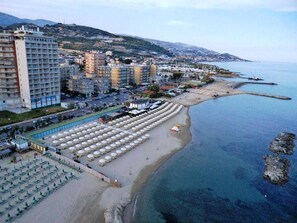A coastal hotel with multiple buildings, a beach with umbrellas, and a view of the sea and hills in the background. - Hotel Svizzera (Taggia)