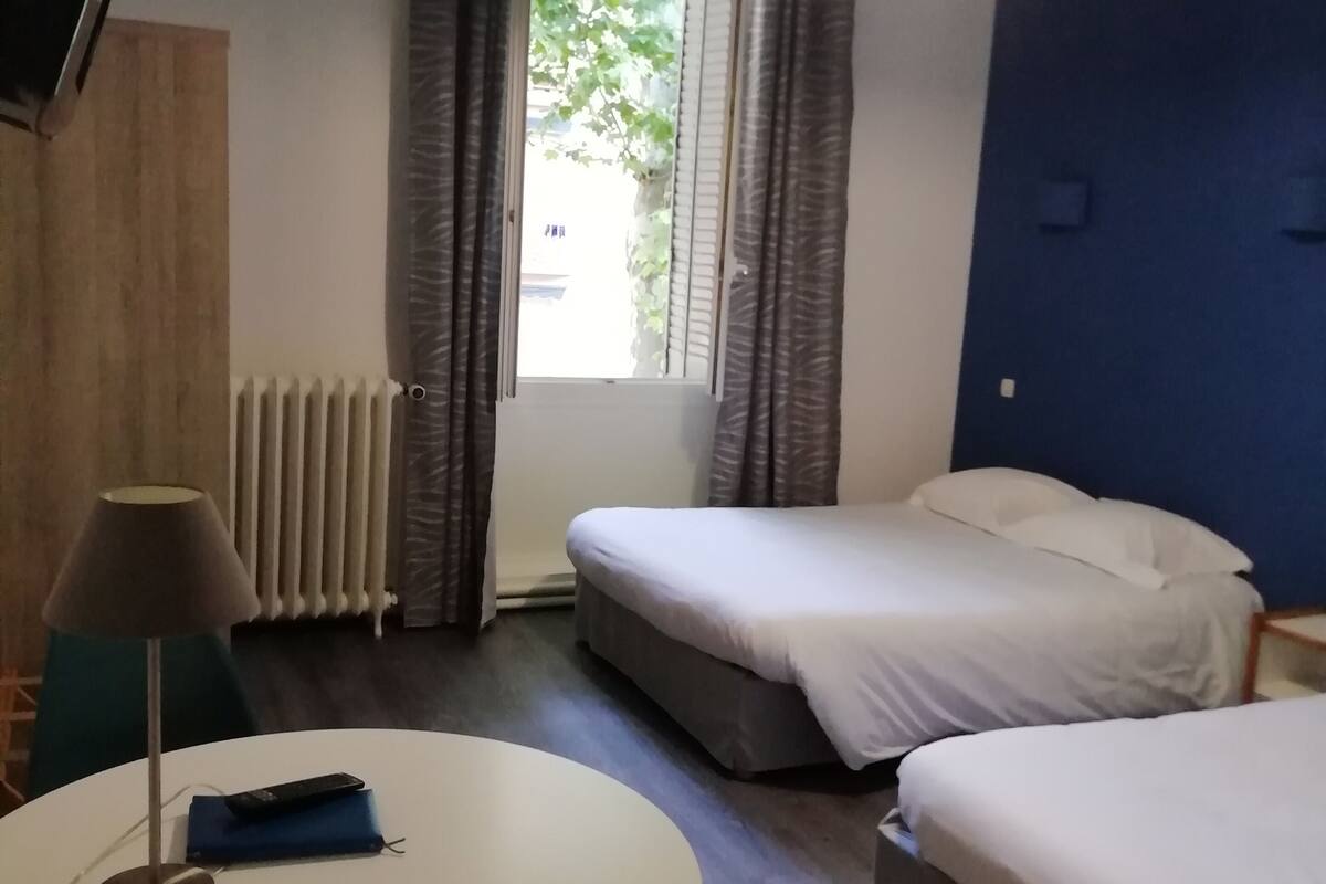 A hotel room with two beds, a round table with a lamp, a radiator, and a window with curtains.